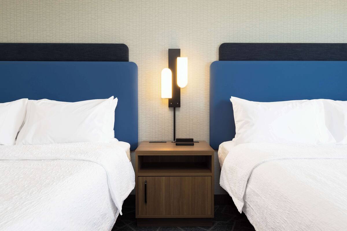 Habitación de hotel con dos camas, una mesita de noche con lámpara y una pared con diseño geométrico.