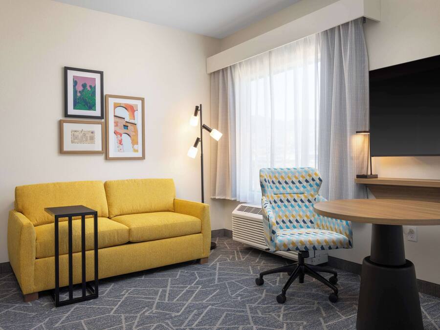 A hotel room with a yellow sofa, a patterned armchair, a small table, and framed artwork on the wall.