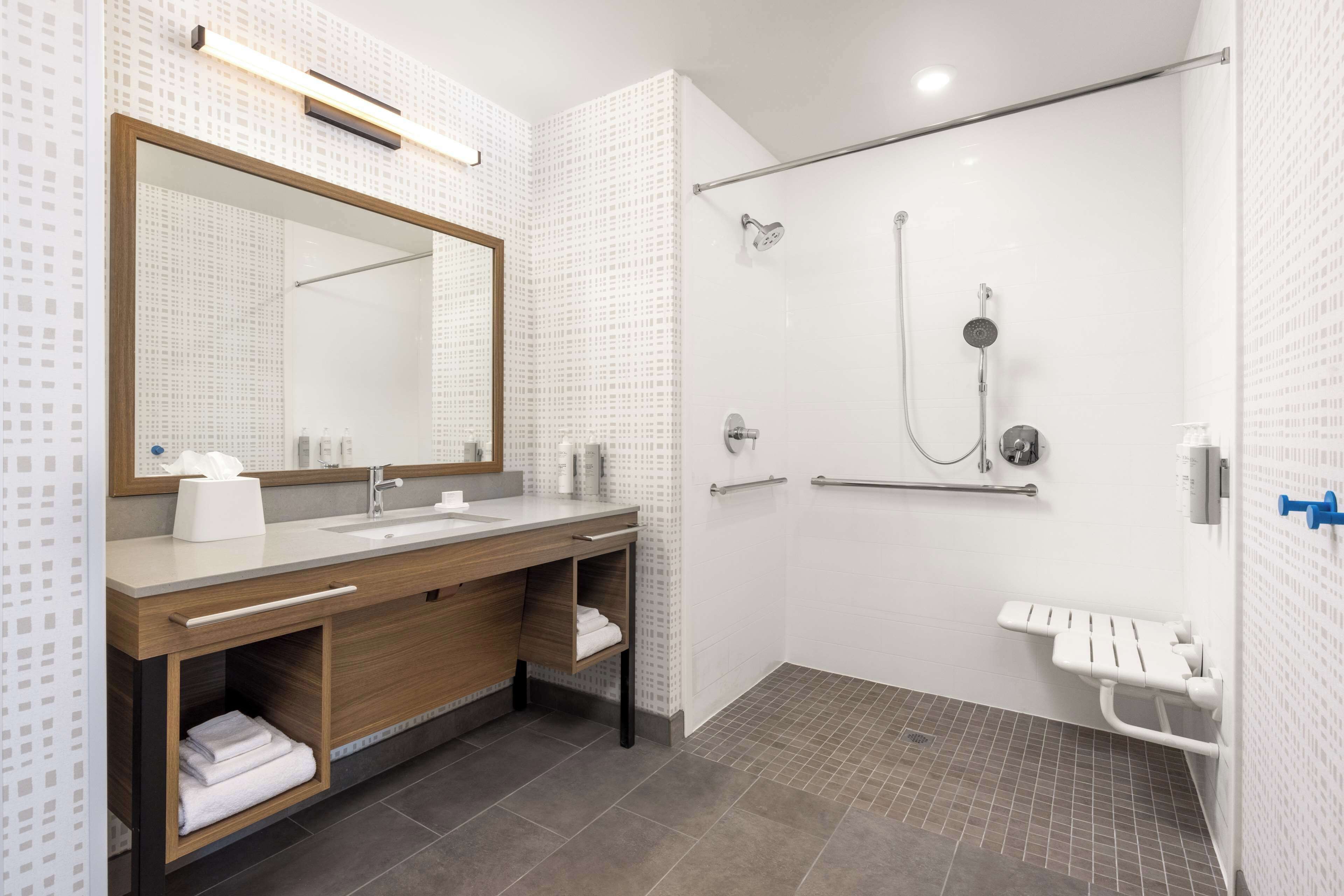 Ein modernes Badezimmer mit einem großen Spiegel, einem Waschbecken, einer Dusche inklusive Handbrause und einer Haltestange.