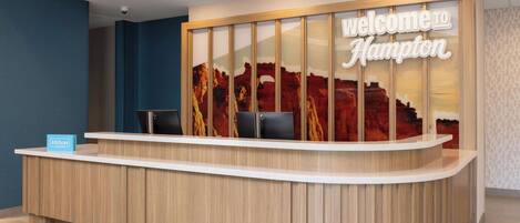 A hotel reception desk with a 'Welcome to Hampton' sign, a blue 'Hilton Honors' sign, and a backdrop of red rock formations.