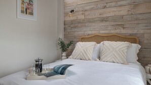 Una cama bien hecha con sábanas blancas, un cojín decorativo y una mesita de noche con un recipiente de vidrio y un libro.