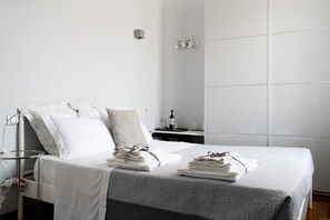 Un letto rifatto con cura, con copriletto grigio fantasia, cuscini bianchi e asciugamani piegati su un comodino.