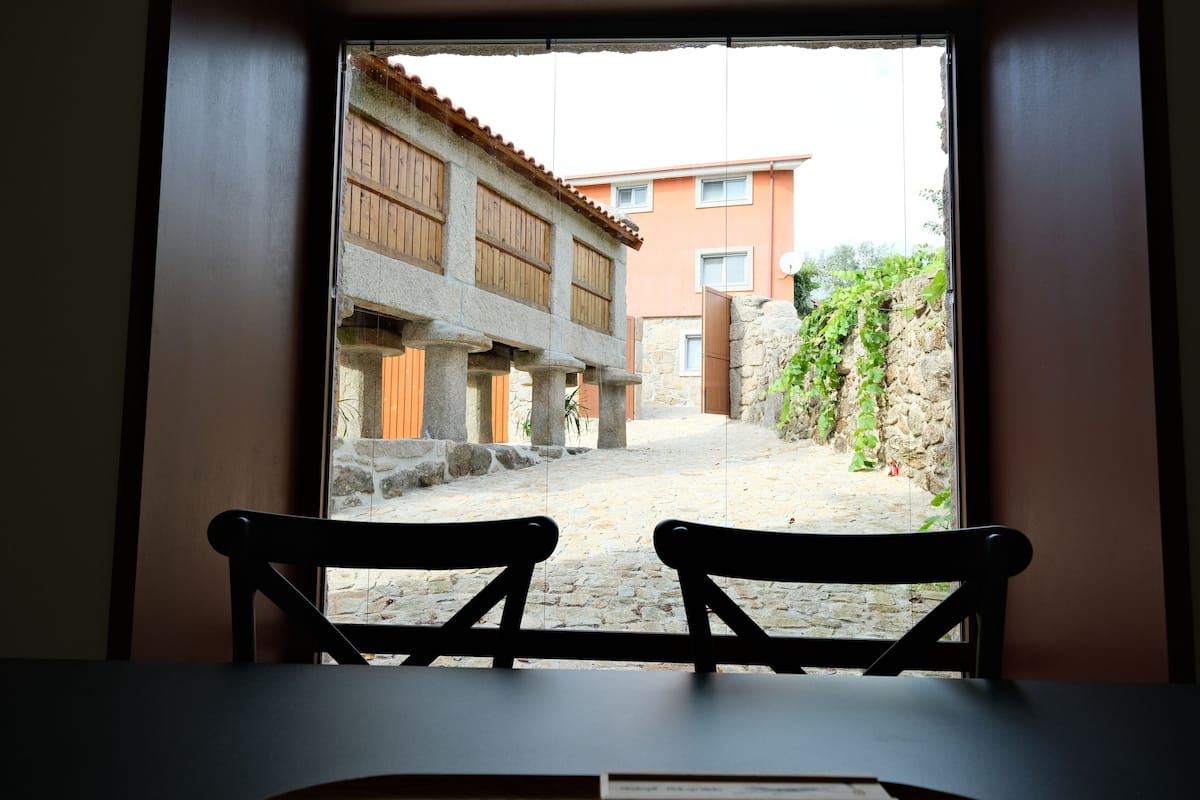 Vista dalla finestra: due sedie di legno, un edificio in pietra con persiane di legno e un cortile lastricato.