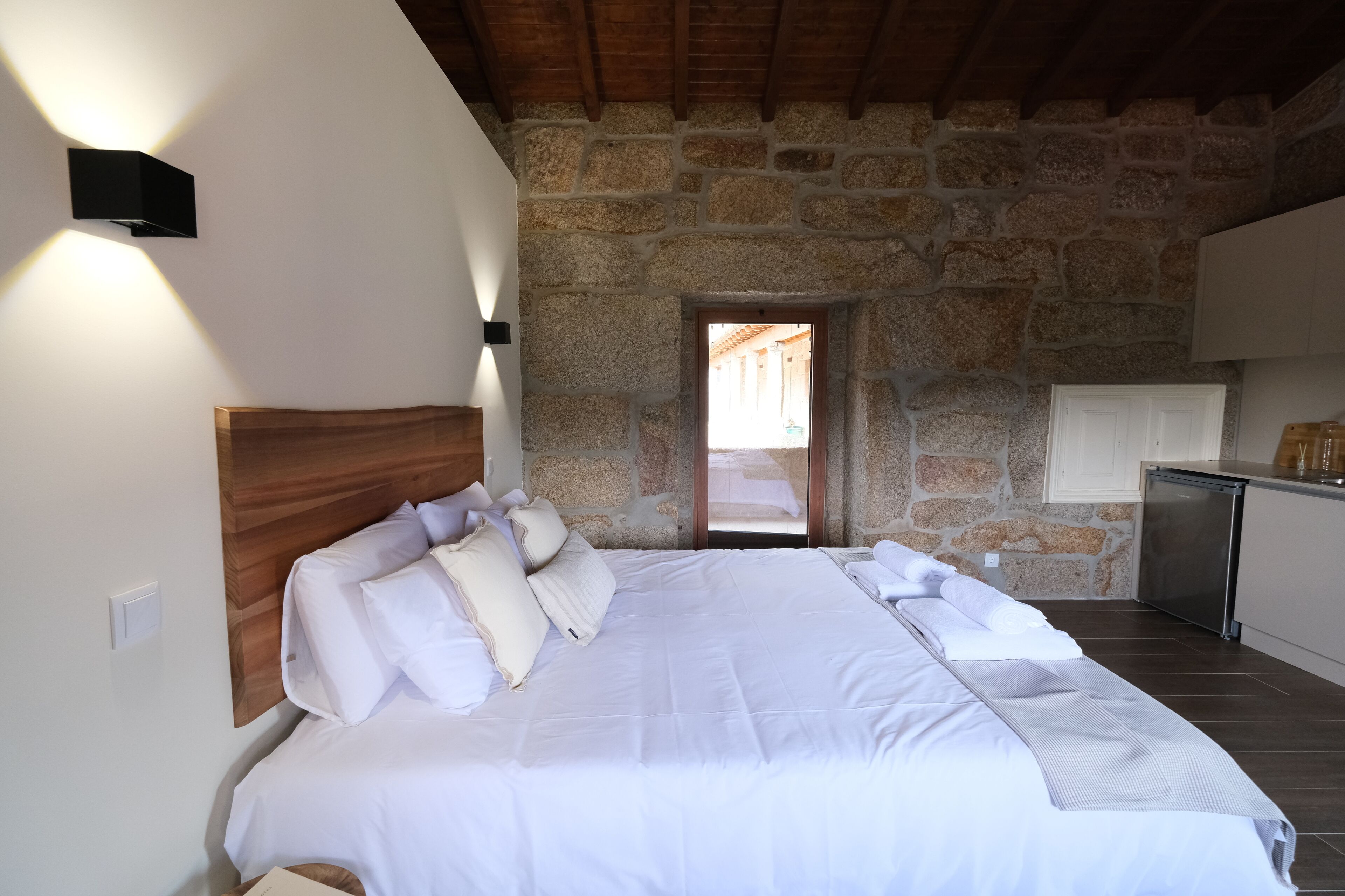 A bedroom with a large bed, stone walls, a wooden ceiling, and a small kitchenette.