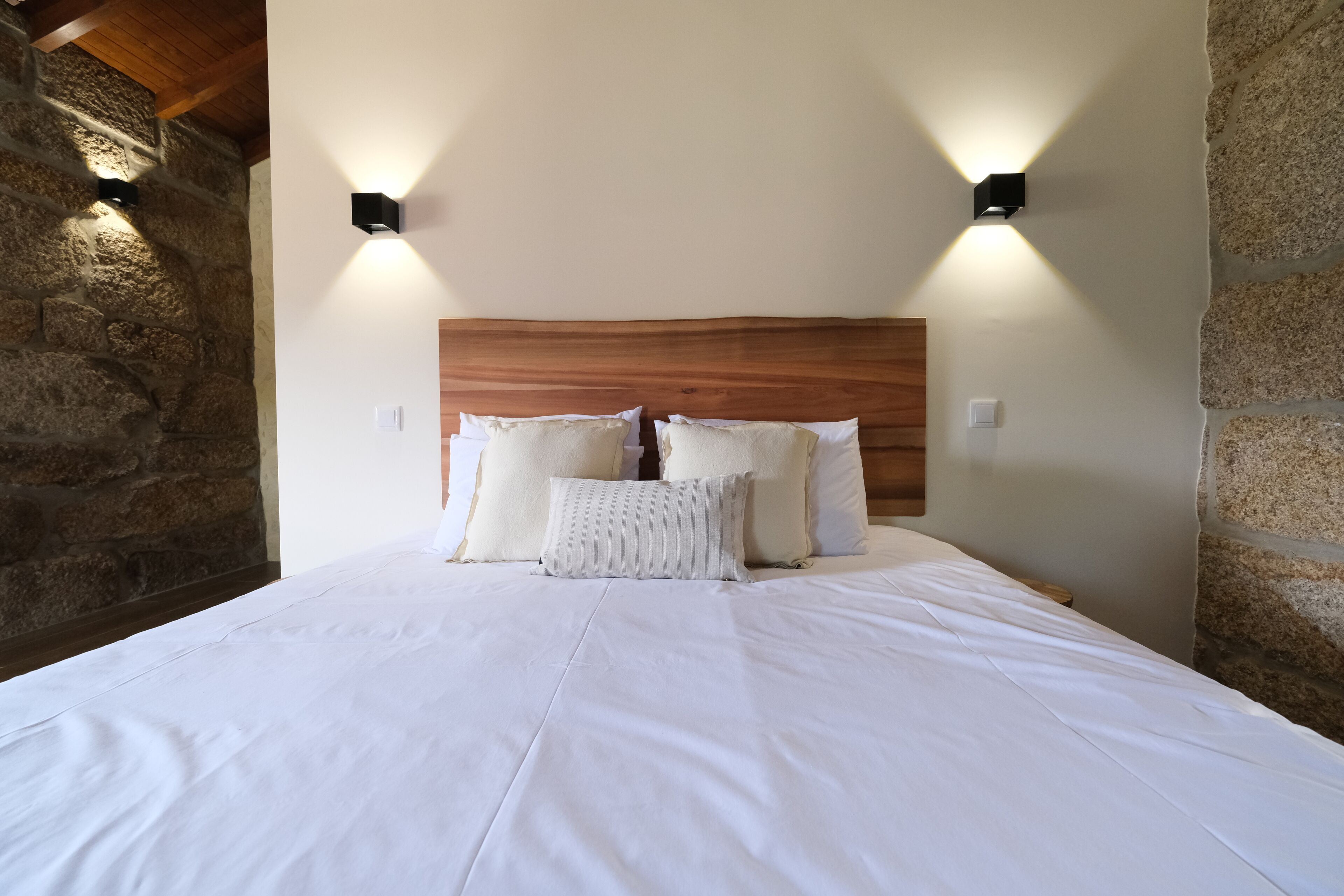 A neatly made bed with white linens and pillows, a wooden headboard, and two wall-mounted light fixtures.