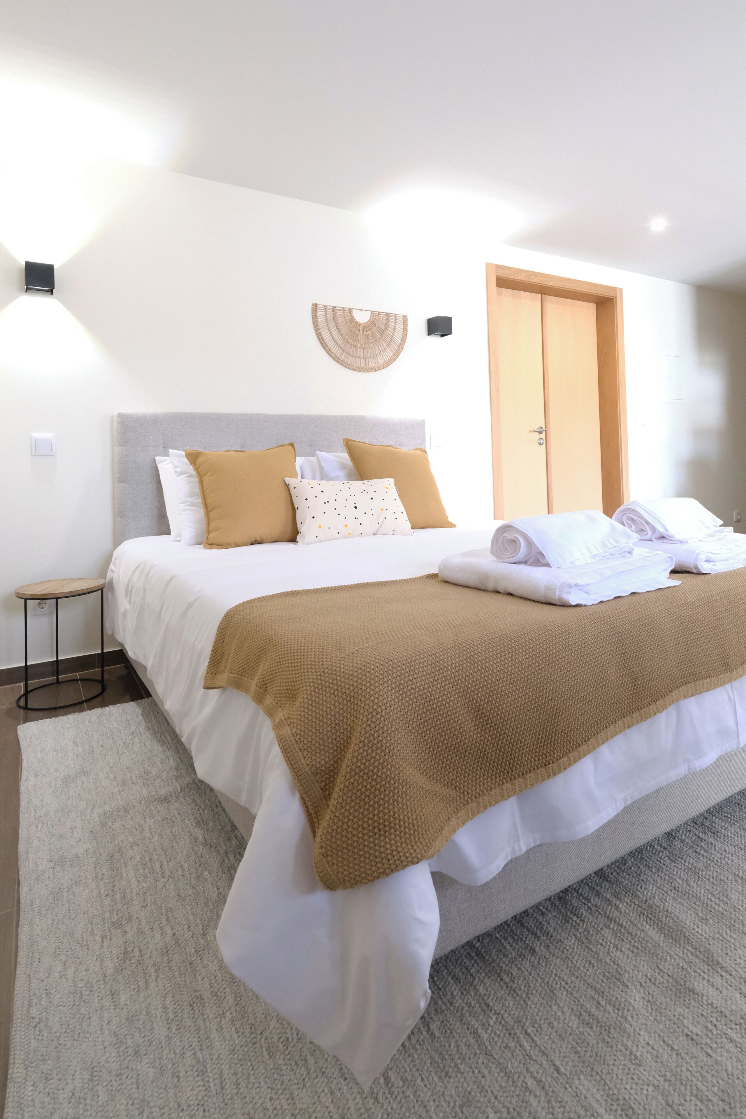 Una cama bien hecha con una manta marrón, una mesita de noche y un ventilador de pared.
