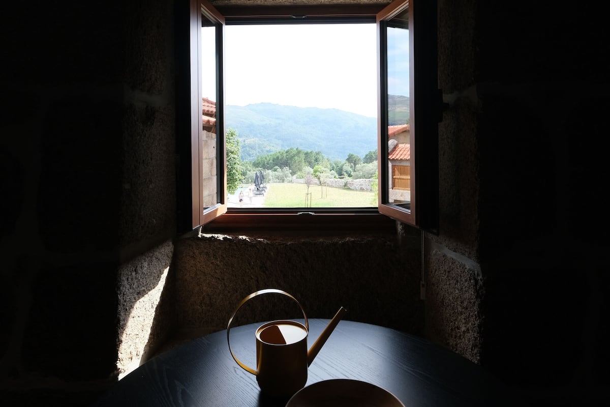 Una stanza scarsamente illuminata con un tavolo e una ciotola. Da una finestra si intravede un paesaggio montuoso.