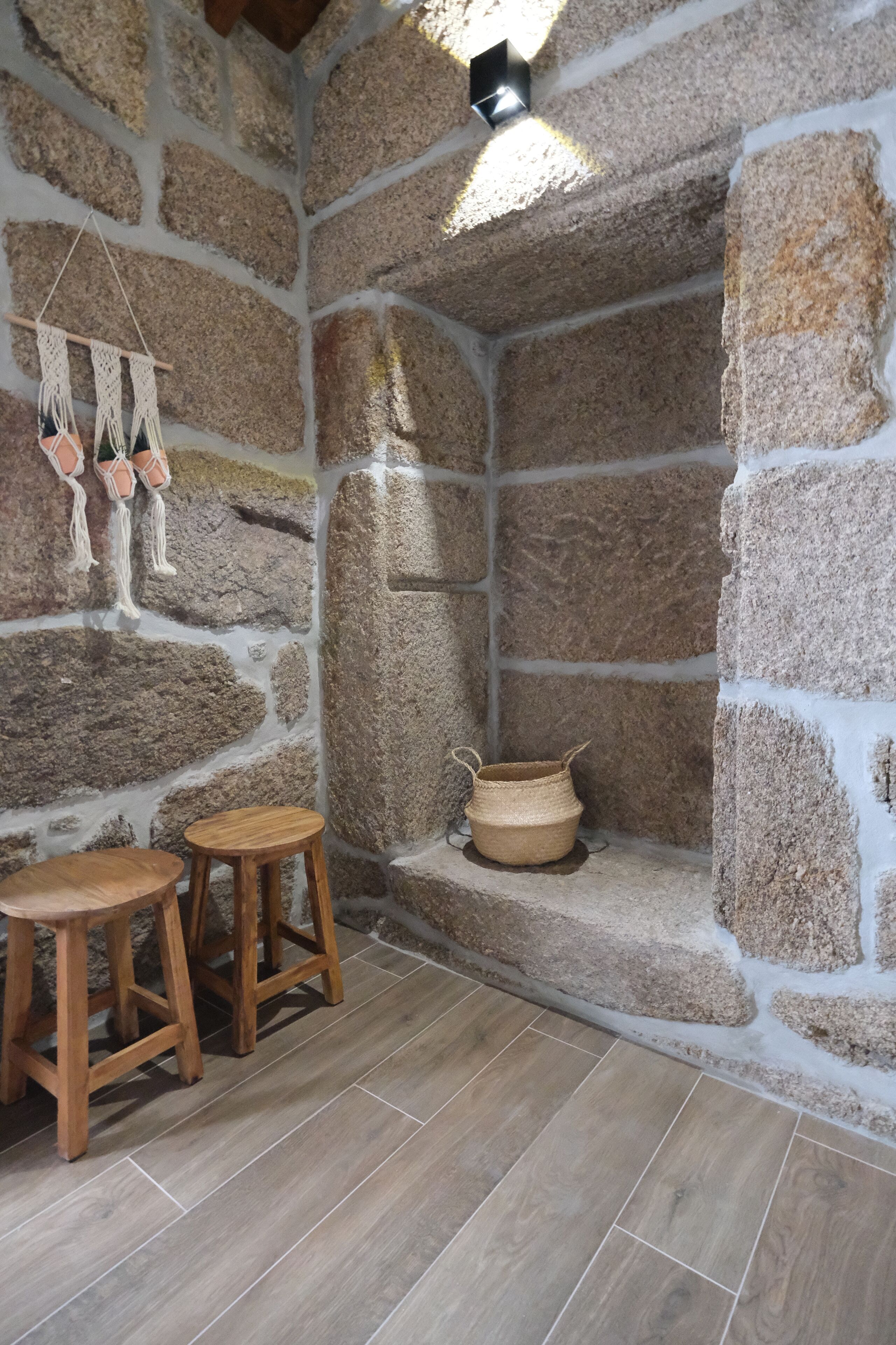 Una habitación con paredes de piedra, un taburete de madera, una decoración de macramé colgante y una cesta tejida en el suelo.