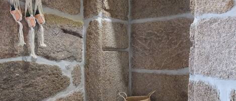 Una habitación con paredes de piedra, un taburete de madera, una decoración de macramé colgante y una cesta tejida en el suelo.