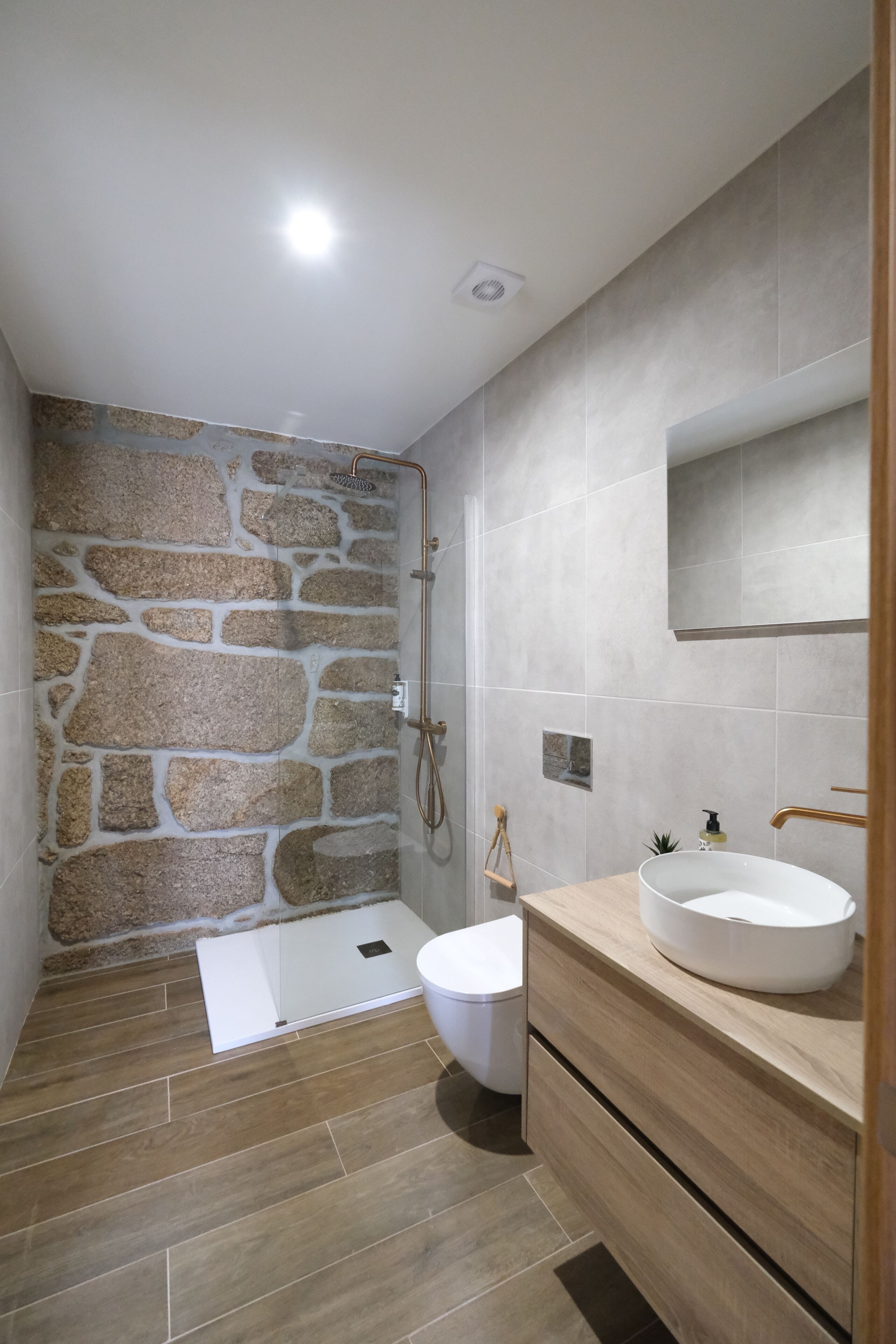A bathroom with a stone wall, a shower, a toilet, and a sink with a mirror above it.