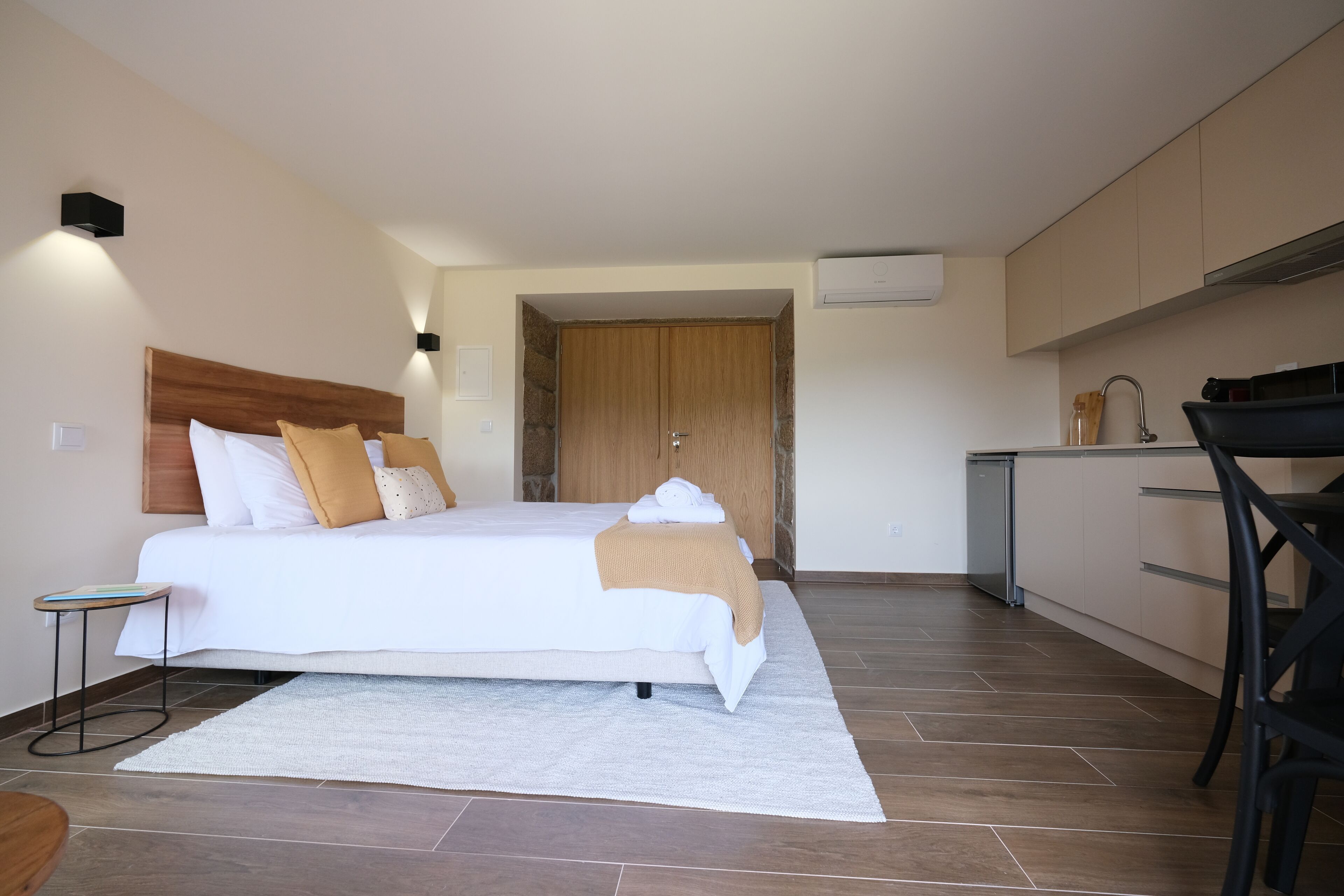 Una habitación de hotel moderna con una cama grande, una mesita de noche pequeña, una zona de cocina con un lavabo y armarios, y una puerta que da a otra habitación.