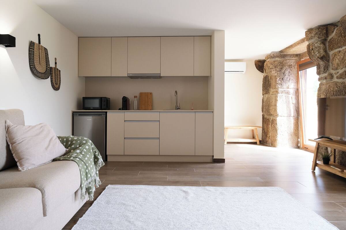 Una cucina moderna con mobili color beige, un forno a microonde e un tagliere di legno. In una zona living adiacente, si nota una parete in pietra e una panca di legno.