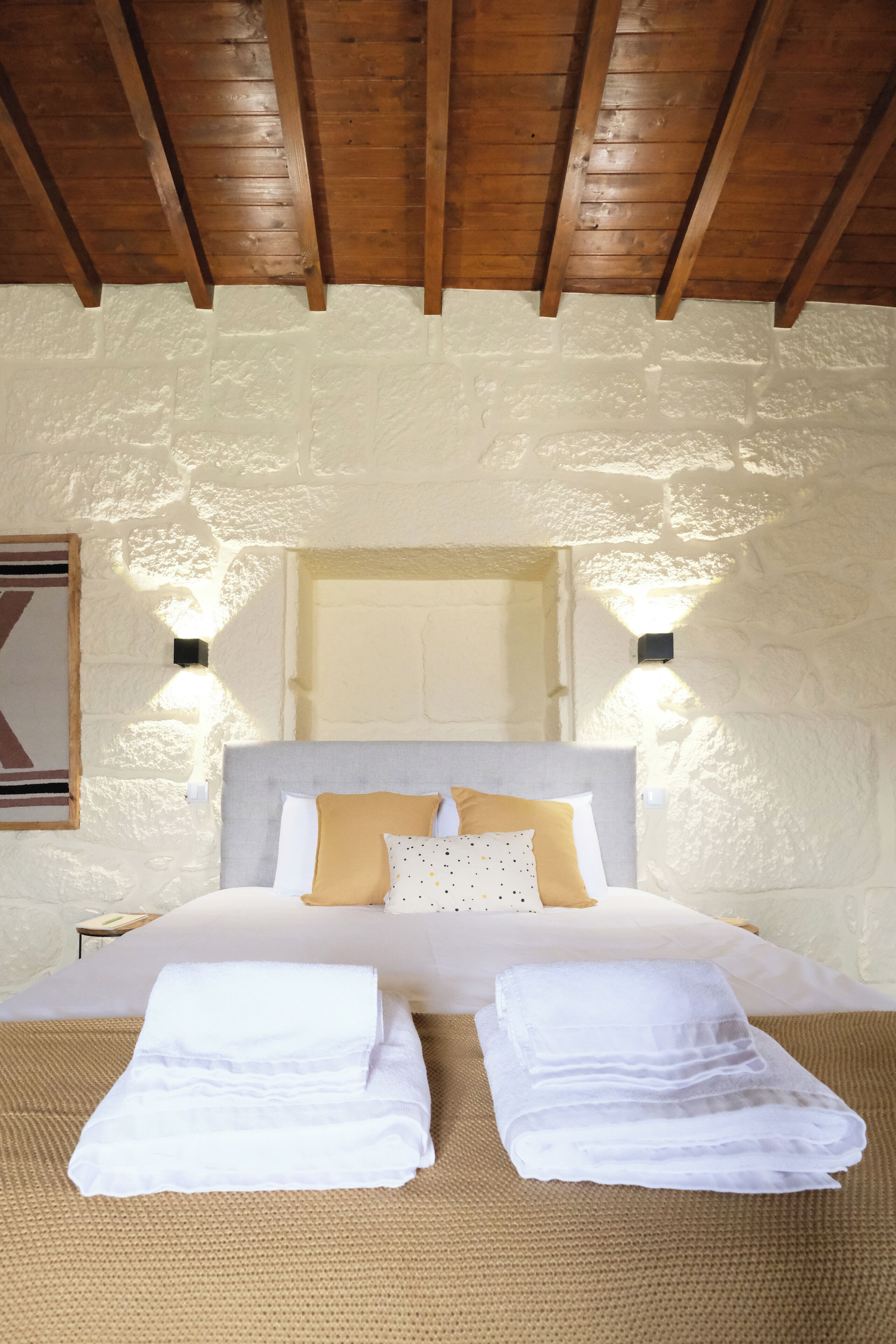 A bedroom with a stone wall, wooden ceiling beams, a bed with white linens, and two wall-mounted lights.