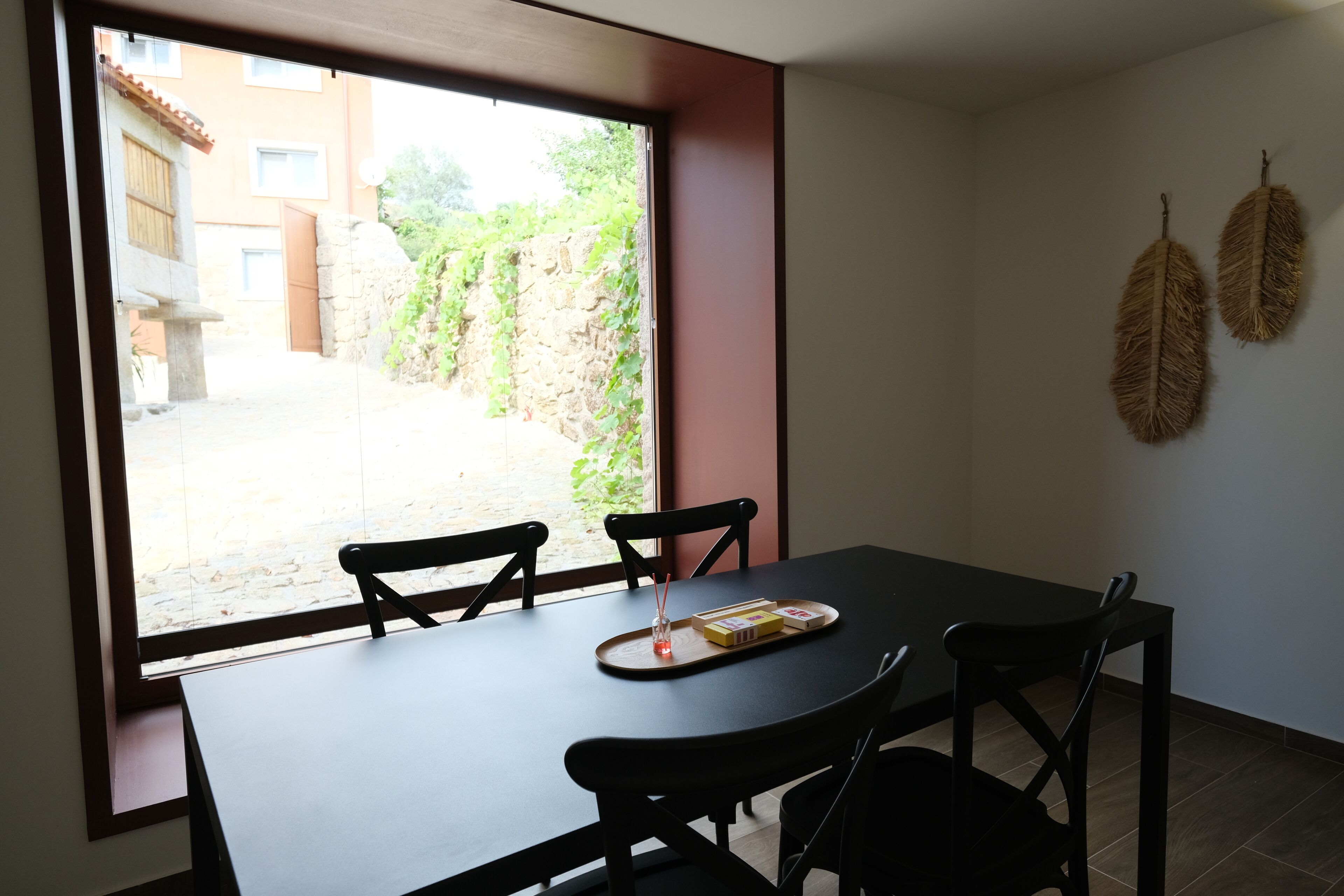 Ein Essbereich mit Tisch und Stühlen, ein Fenster mit Blick auf einen Innenhof und zwei an der Wand befestigte dekorative Blätter.