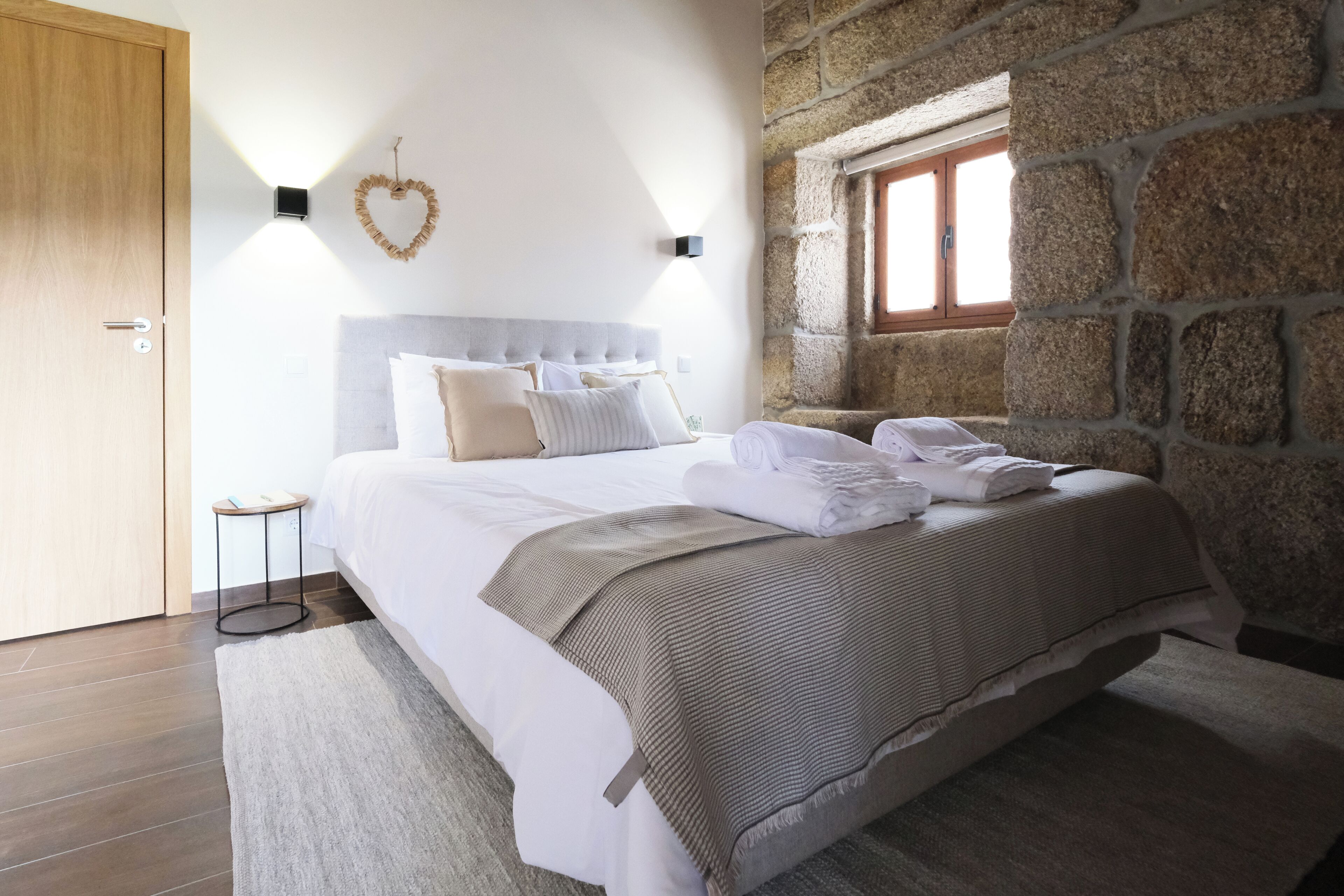 A bedroom with a large bed, a heart-shaped wall decoration, a stone wall, and a window.