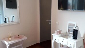 Une pièce comprenant une coiffeuse blanche, un miroir, un téléviseur fixé au mur, ainsi qu’une petite table blanche sur laquelle se trouvent une bouilloire et des tasses.