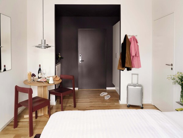 A hotel room with a bed, a desk with a chair, a small table with snacks and drinks, a coat rack with jackets, a suitcase, and a door leading to another room.