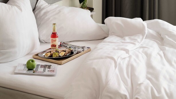 Ein ordentlich bezogenes Bett mit weißer Bettwäsche, eine Flasche Fern Wein, ein Tablett mit Speisen, ein grüner Apfel und eine Zeitung auf einem Holztablett.