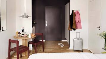 Ein Hotelzimmer mit Bett, Schreibtisch mit Stuhl, einem kleinen Tisch mit Snacks und Getränken, einem Kleiderständer mit Jacken, einem Koffer und einer Tür zu einem weiteren Raum.