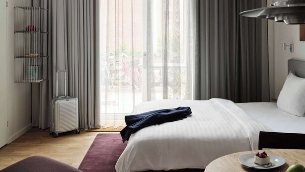 A hotel room with a bed, a desk with a plate of fruit, a chair, and a window with curtains. - Studio for 2 with High Ceilings (København)