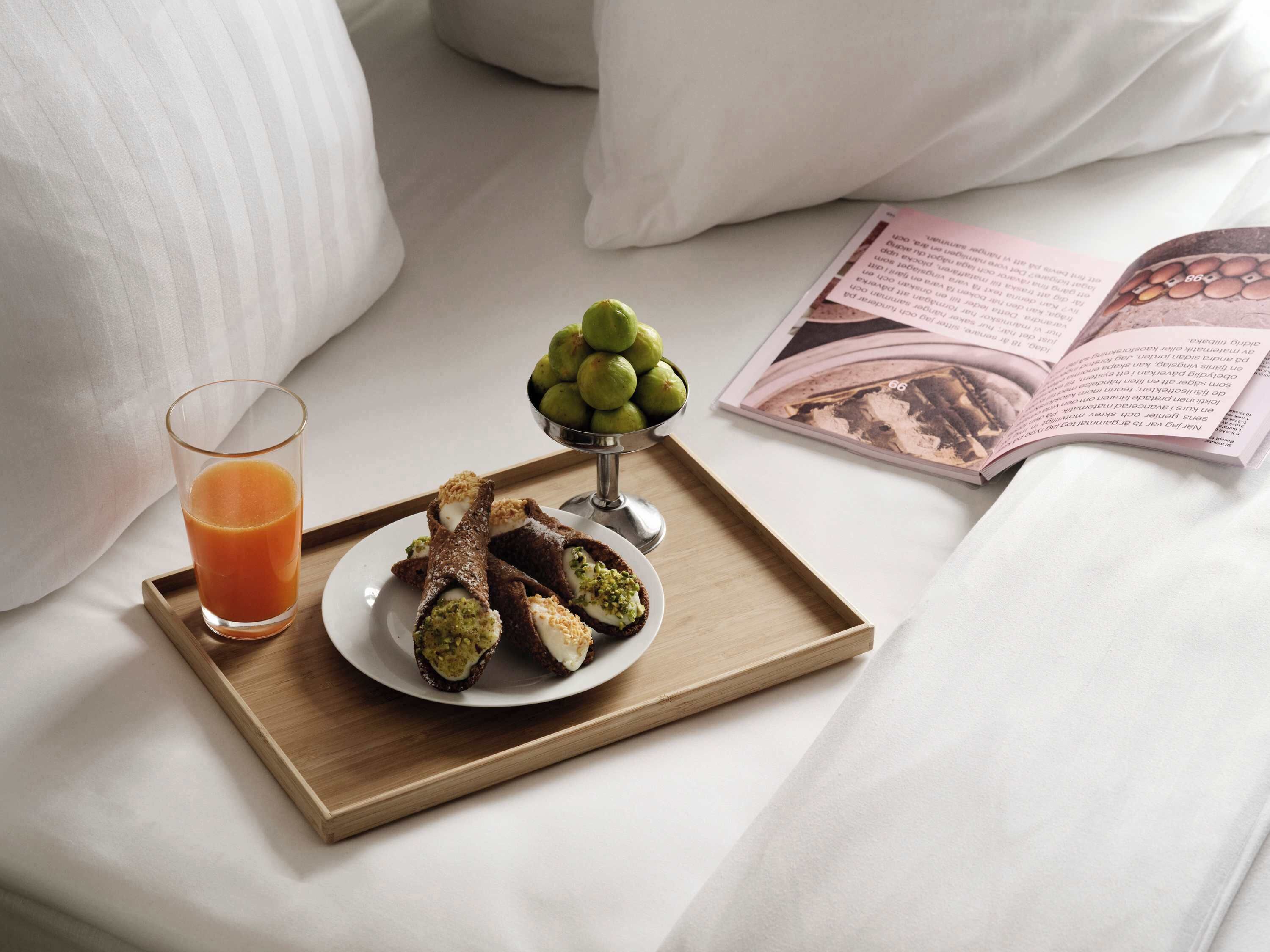 A tray with a glass of orange juice, a plate of pastries, and a bowl of green fruits on a bed with pillows and an open book.