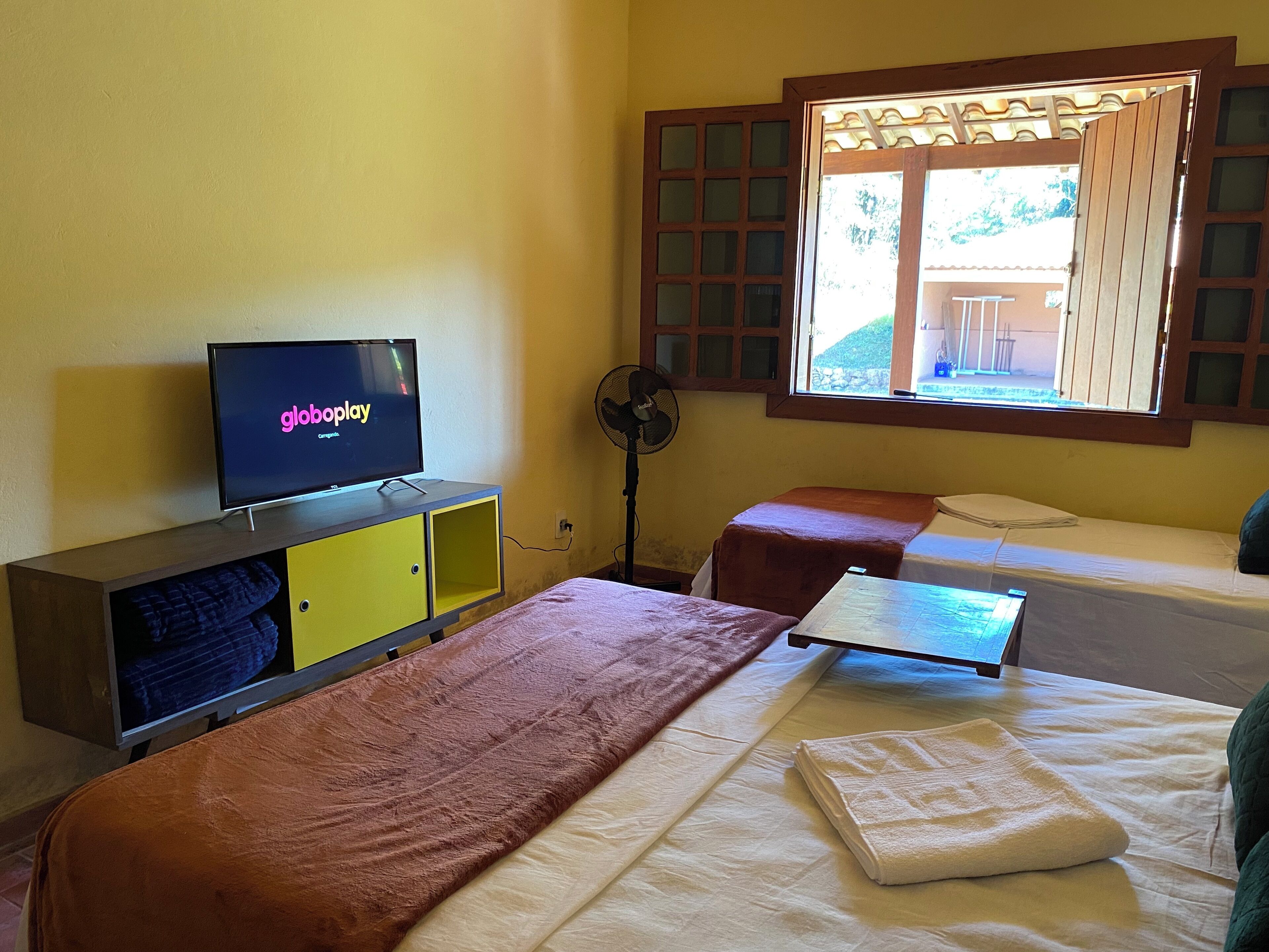 A room with two beds, a TV displaying 'globalplay', a fan, and a window with a view of greenery.