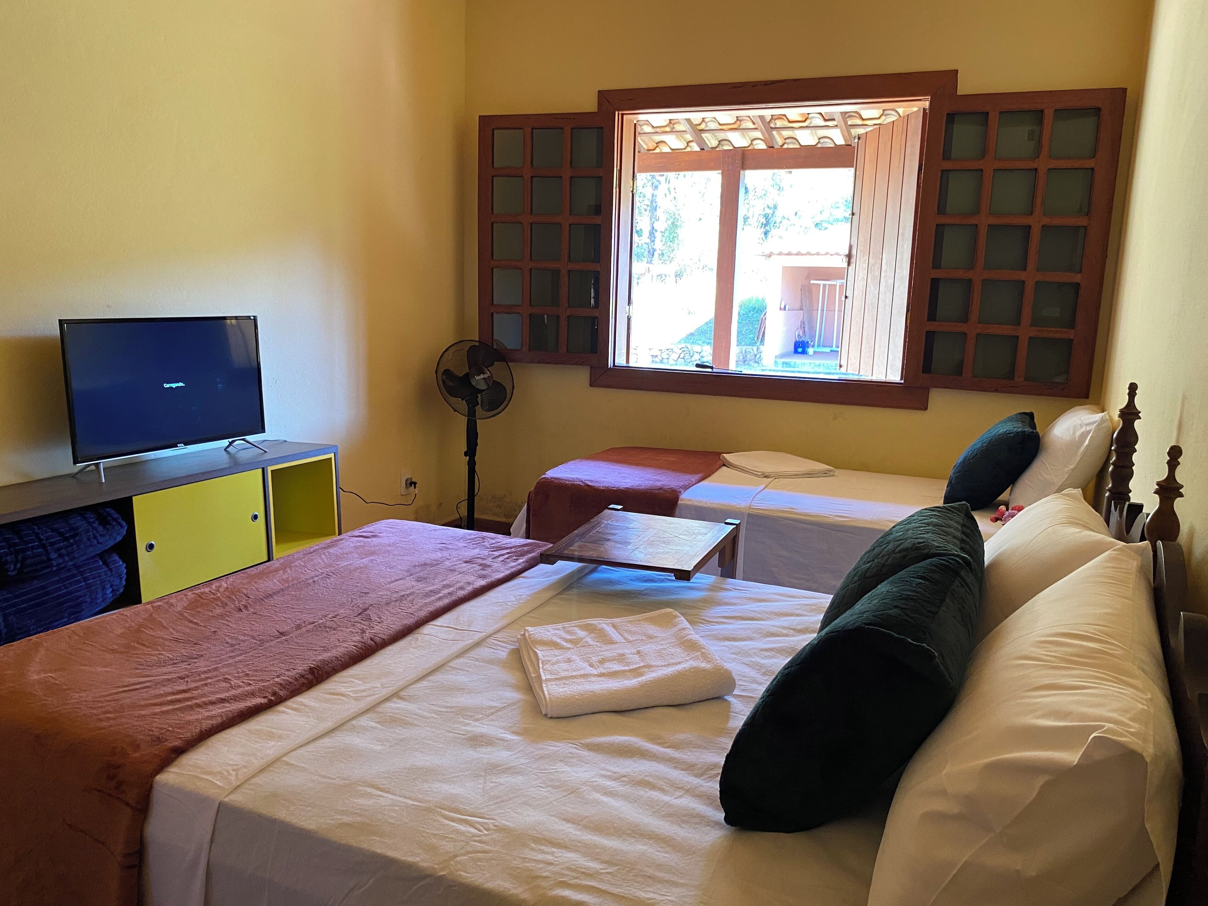 Quarto de hotel com duas camas de solteiro, uma TV sobre um armário de madeira, um ventilador e uma janela com venezianas de madeira.
