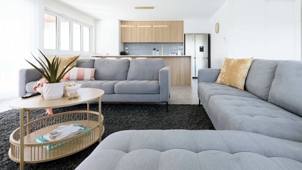 A modern living room with a grey sofa, a coffee table, and a kitchen in the background. - Spacious 4-Bed Coastal Retreat with Ocean Views (Bridport)