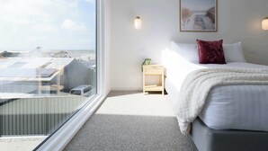 A hotel room with a large window, a bed with white bedding, a red pillow, a small bedside table with a plant, and a framed picture on the wall. - Spacious 4-Bed Coastal Retreat with Ocean Views (Bridport)