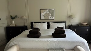A neatly made bed with two folded towels, a bedside table with a lamp, a framed abstract artwork, and a small plant on the nightstand. - Cozy Manchester Hideaway With 2 Bedrooms (Manchester)