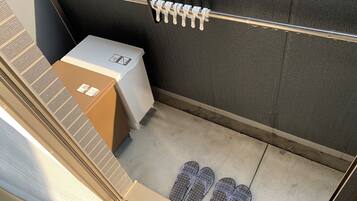 A small bathroom with a shower, a toilet, and a trash bin. There are slippers on the floor and a towel rack with a towel hanging on it.