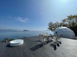 A wooden deck with a hot tub, a table with chairs, and a covered seating area overlooking a body of water.