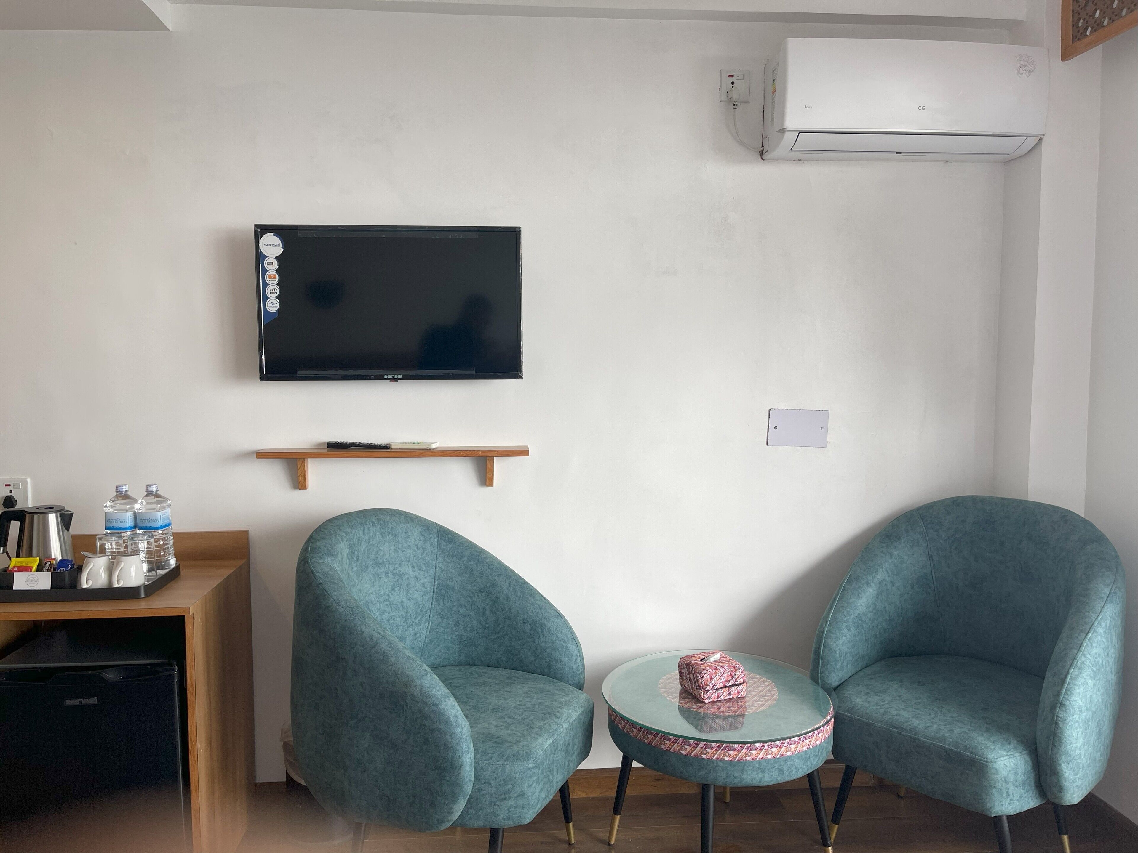 A modern living room with a flat-screen TV, two teal armchairs, a small round table, and a coffee station.
