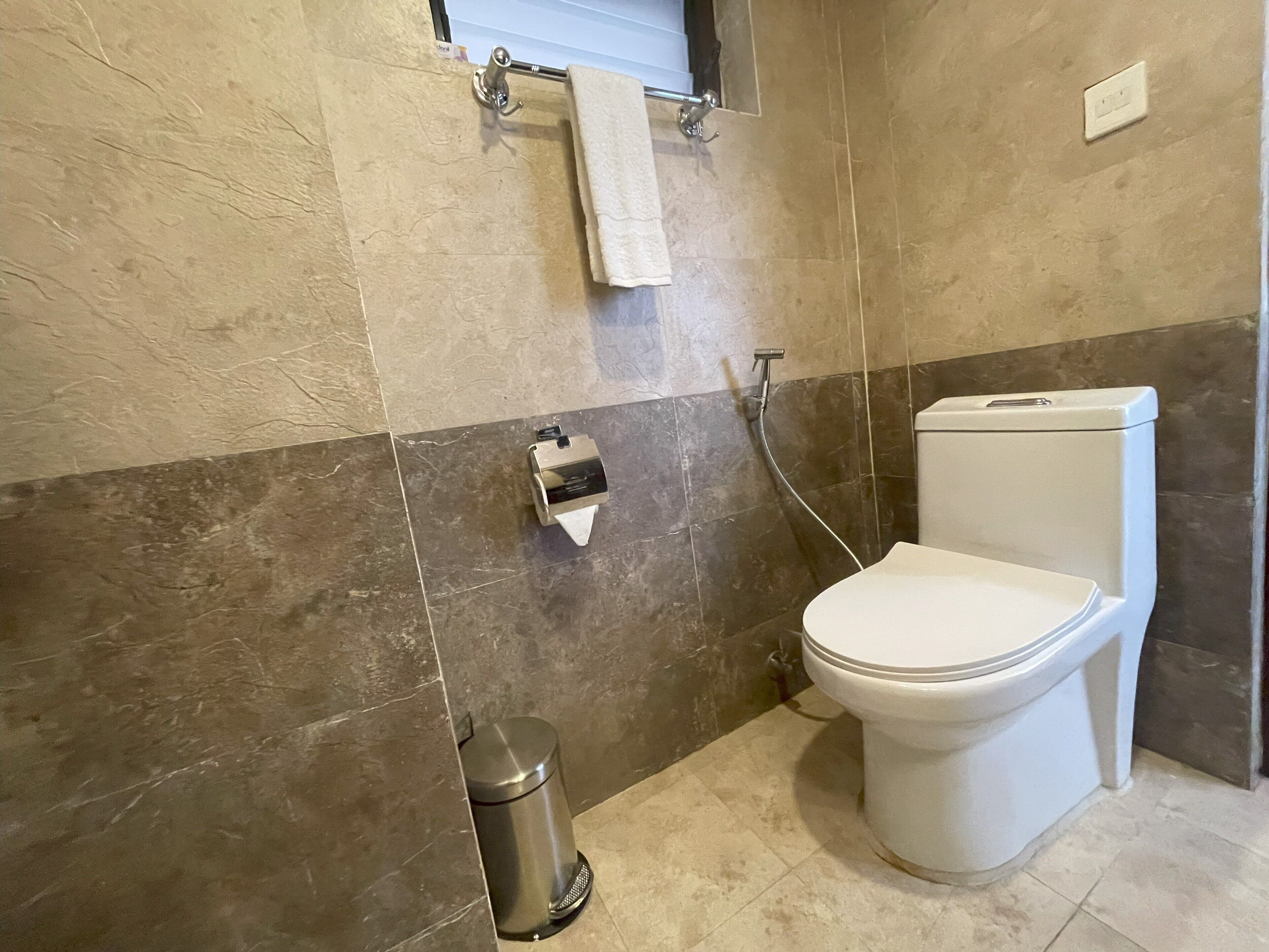 A bathroom with a toilet, a trash bin, a towel rack, and a shower head.