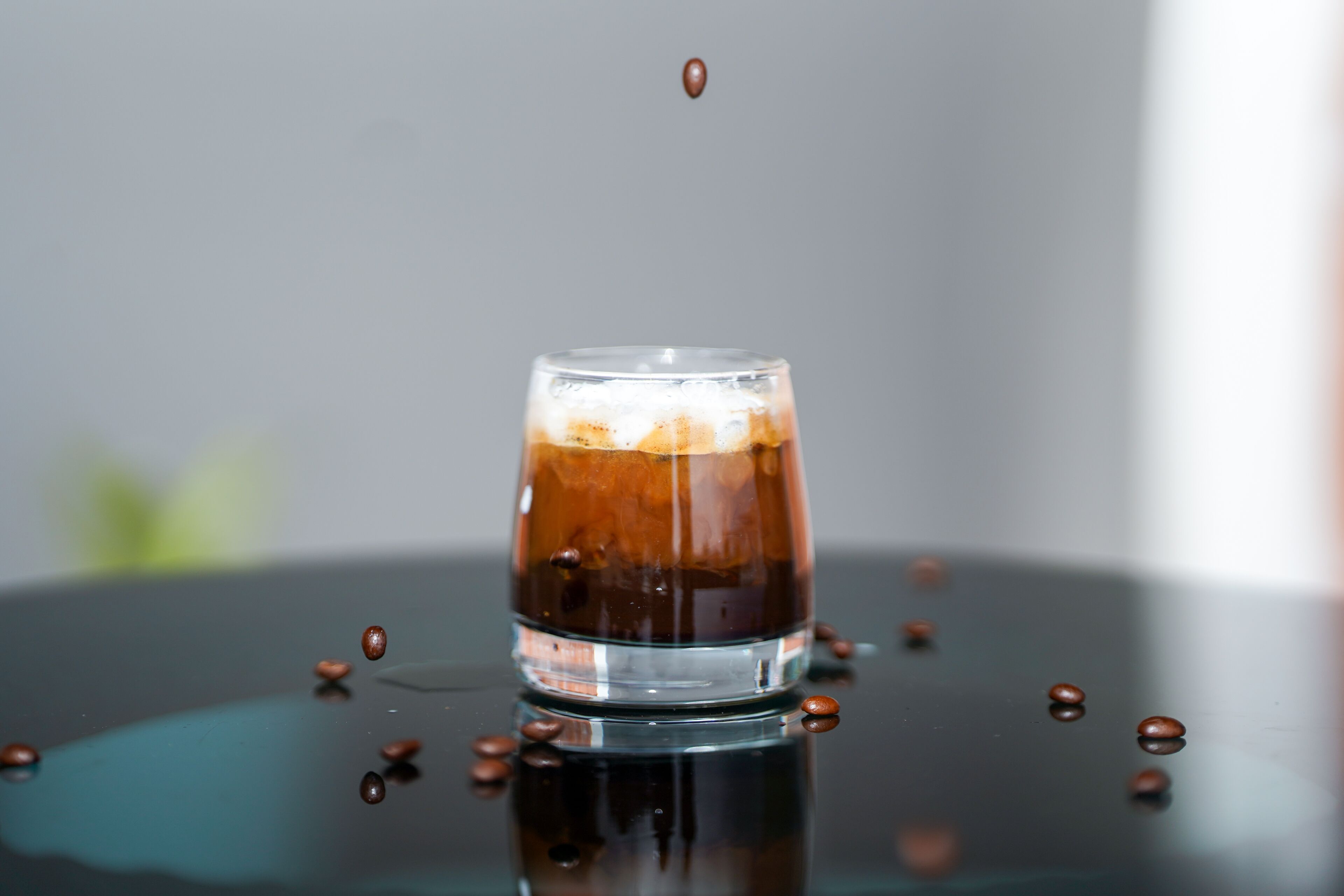 A glass of iced coffee with floating coffee beans on a reflective surface.