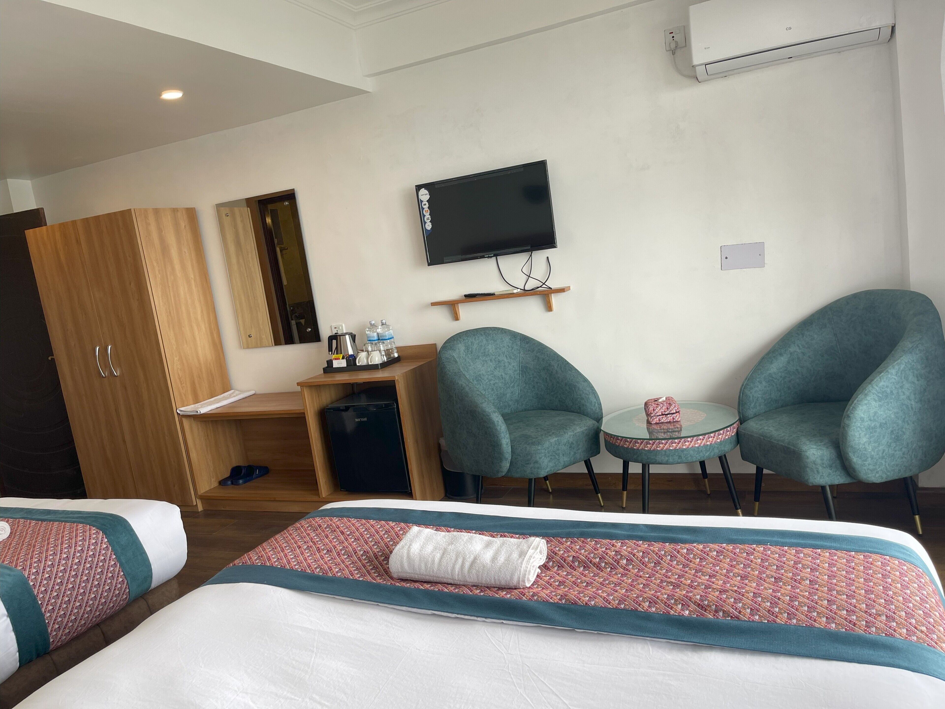 Habitación de hotel con dos camas individuales, un escritorio con silla, un televisor y una pequeña mesa con una silla.