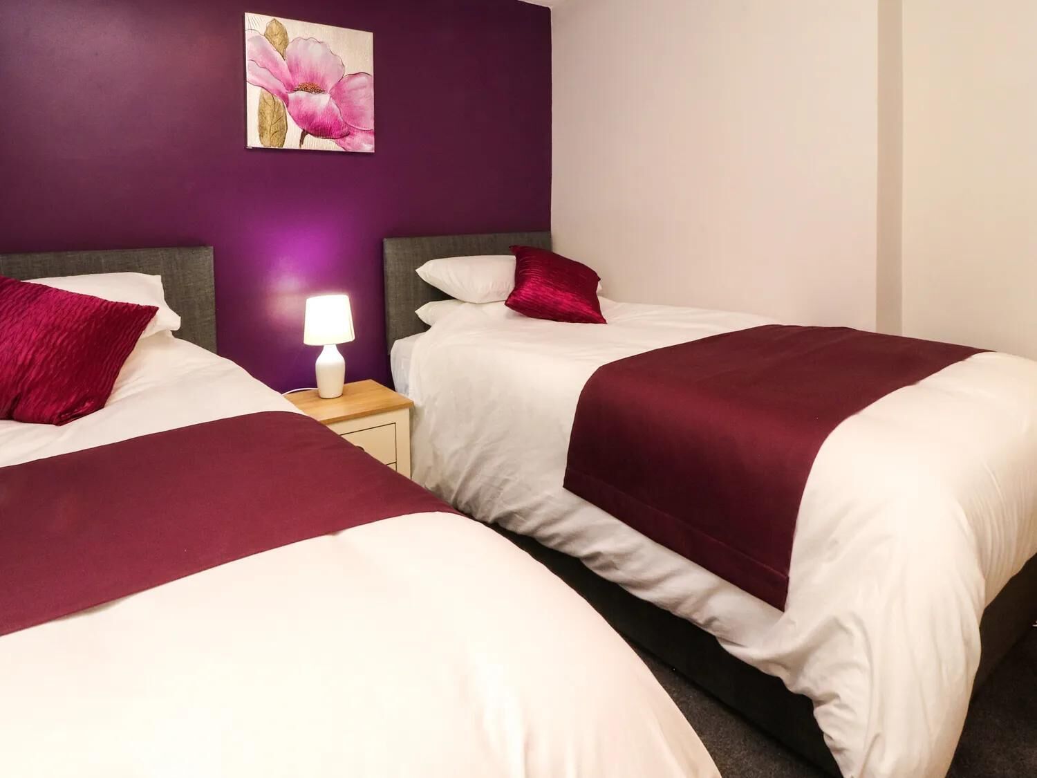 A hotel room with two beds, each with white bedding and a maroon throw blanket. There is a nightstand with a lamp between the beds.