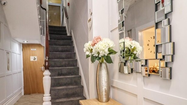 A staircase with a wooden handrail and carpeted steps leading to a landing. On the landing, there is a wooden console table with a decorative plate and a vase with flowers. - 7-bed Beachside Retreat - Sleeps 14 & Pet Friendly (Blackpool)