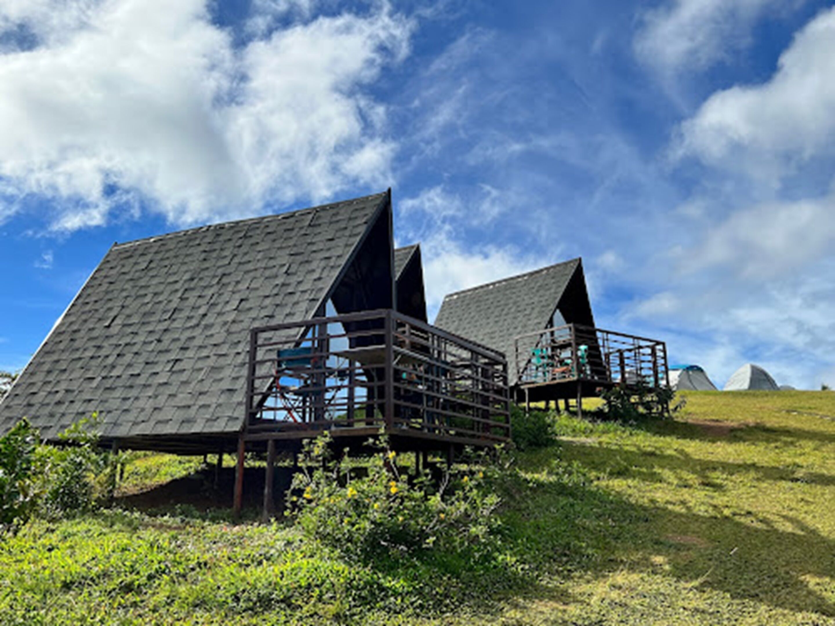 Een rij houten vakantiehuisjes met puntige daken op een groene heuvel, onder een heldere lucht.