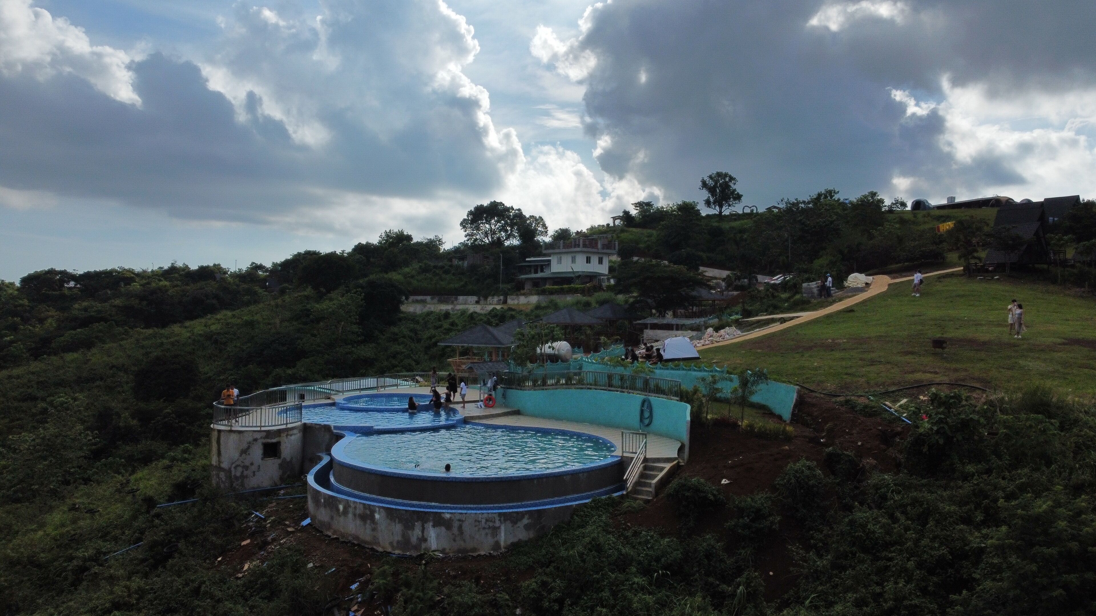 A swimming pool complex with multiple circular pools, surrounded by greenery and a few buildings in the background.