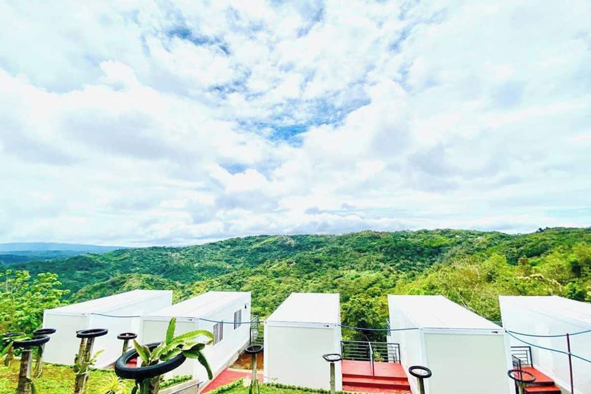 A row of white cabins with red roofs, surrounded by lush greenery and a mountainous backdrop.