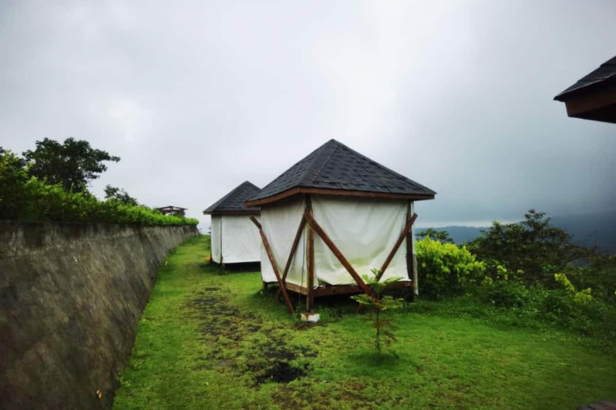 A row of simple, white tents with dark roofs, set on a grassy path beside a stone wall, with a cloudy sky above.