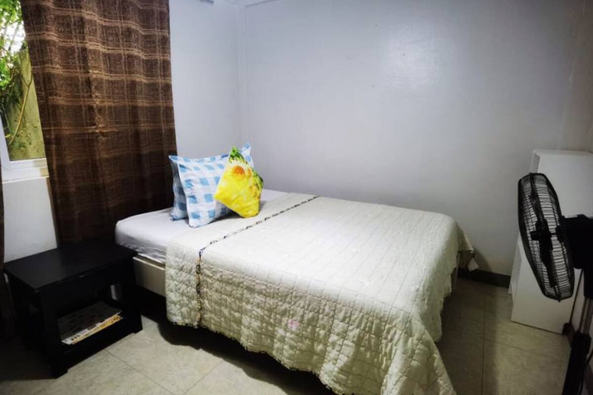 A small, clean room with a bed, a nightstand, a fan, and a window with curtains.