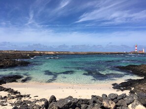 A beach with clear turquoise water, a sandy shore, and a lighthouse in the distance.