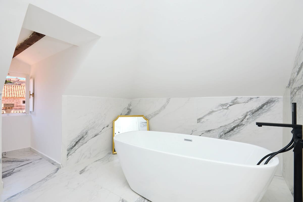 A modern bathroom with a freestanding bathtub, marble walls, and a small window.