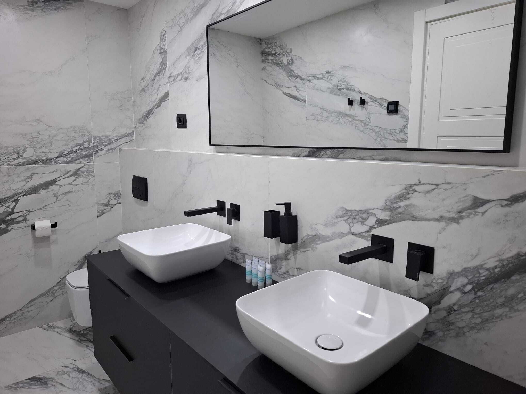 Un baño moderno con dos lavamanos blancos, dispensadores de toallas negros y un espejo grande arriba.