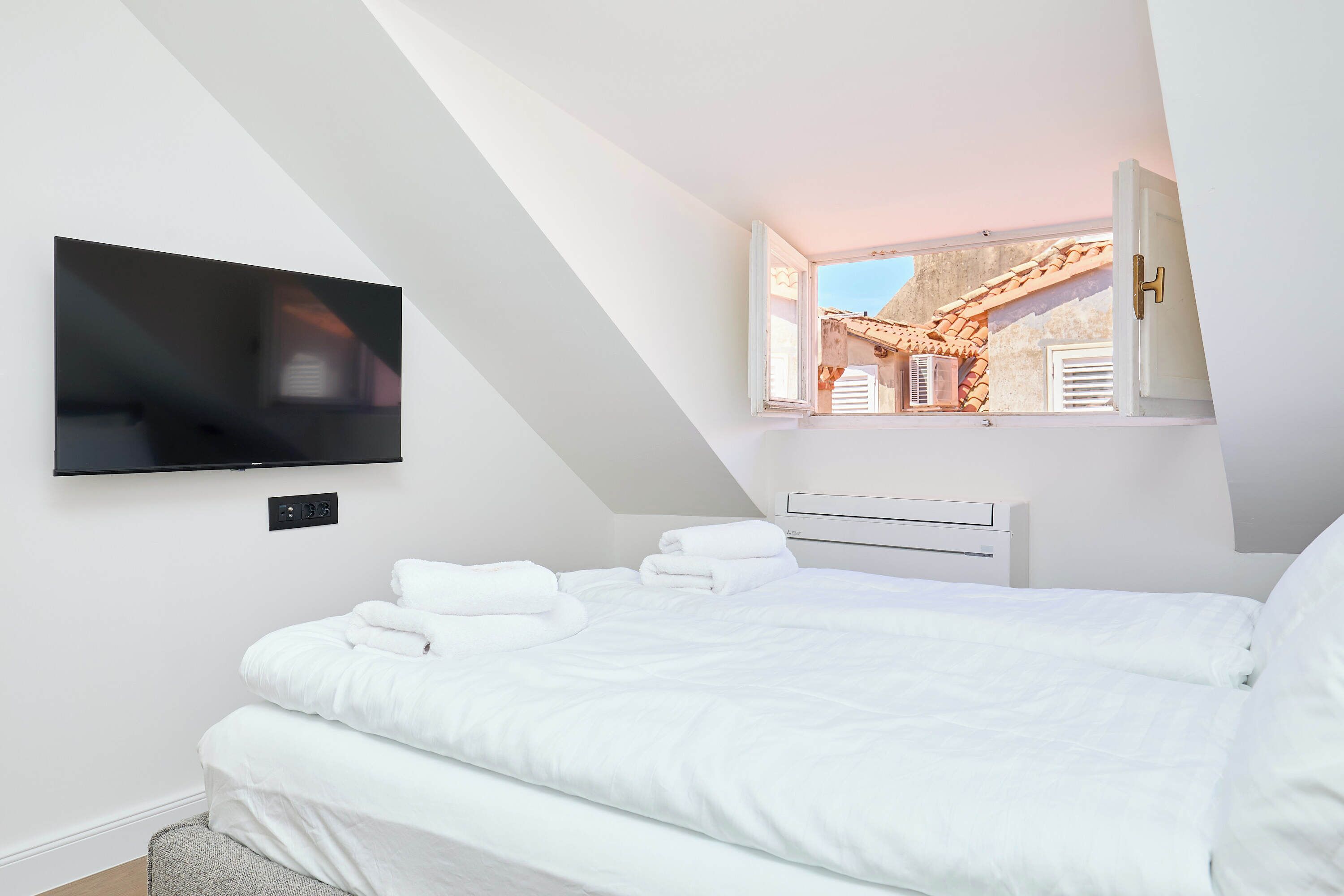 A modern bedroom with a bed, a flat-screen TV mounted on the wall, and a window with a view of a building.