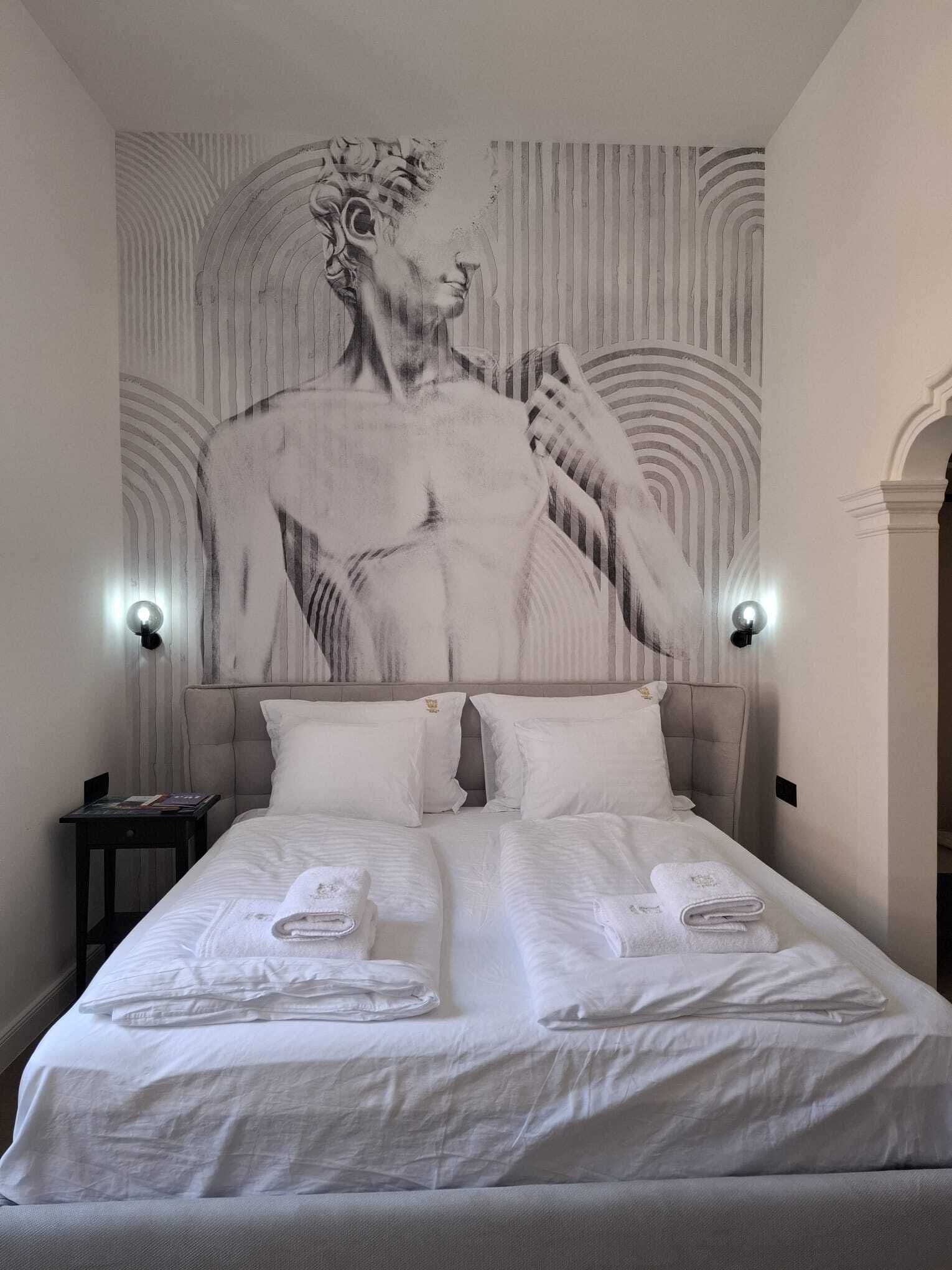Habitación de hotel con un gran mural de una escultura clásica en la pared, una cama con ropa blanca y dos lámparas de noche.