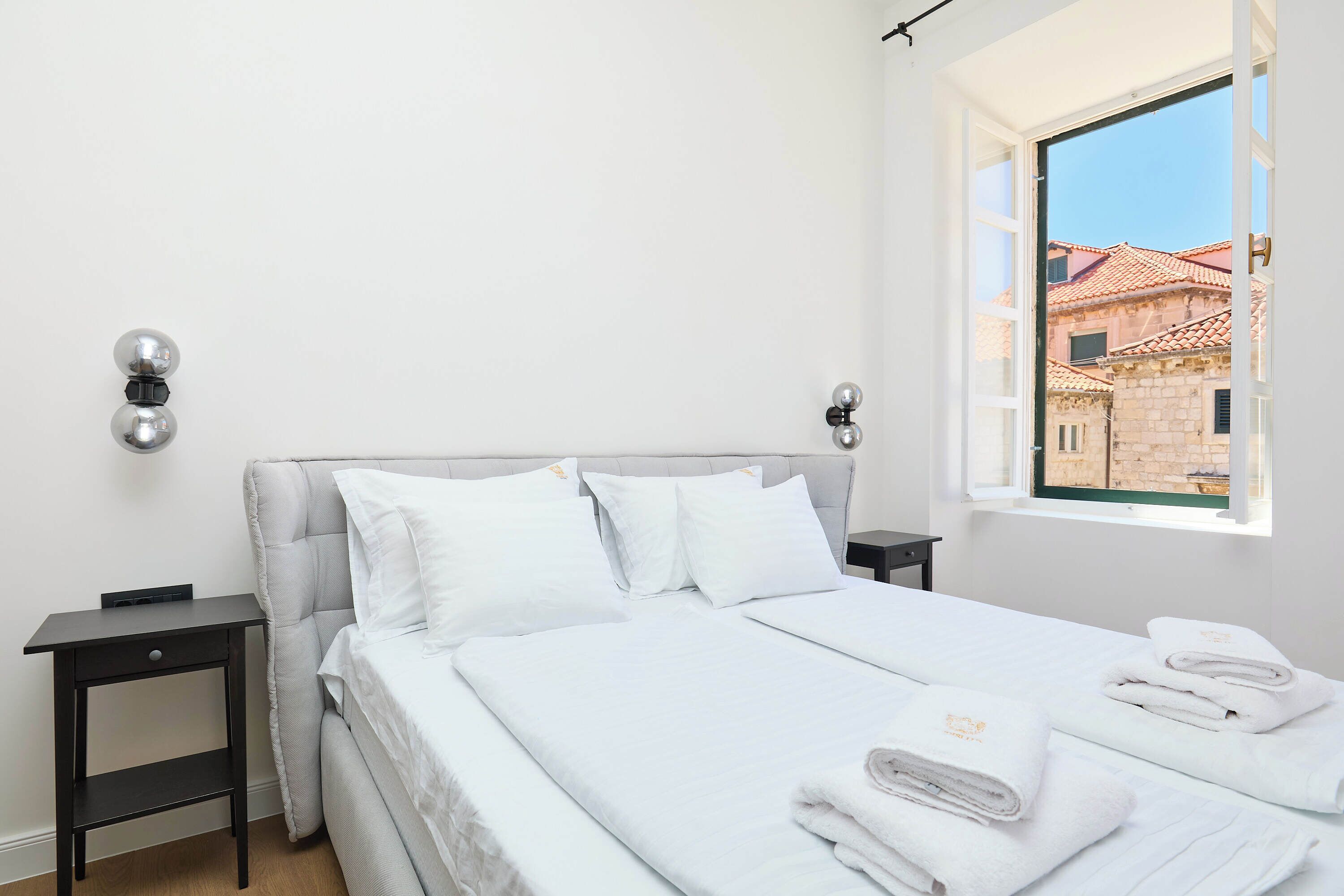 Cama bem arrumada com lençóis e almofadas brancas, uma pequena mesa de cabeceira com abajur e uma janela com vista para prédios.