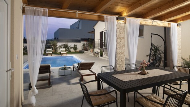 Un patio al aire libre con piscina, techo de madera, cortinas blancas y una zona de comedor con sillas y una mesa.